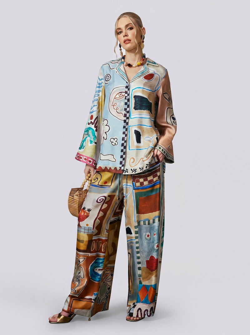 A woman stands against a plain background, wearing the Social Queen Abstract Print Mulberry Silk Shirt with a loose resort-wear vibe, its colorful painterly design making a statement. She completes her look with a small wicker handbag, earrings, necklace, and sandals.
