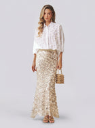 Dressed in Social Queen’s Pearl-Embellished Floral Blouse and a long, sequined beige skirt, the woman holds a small handbag while standing against a plain light background.