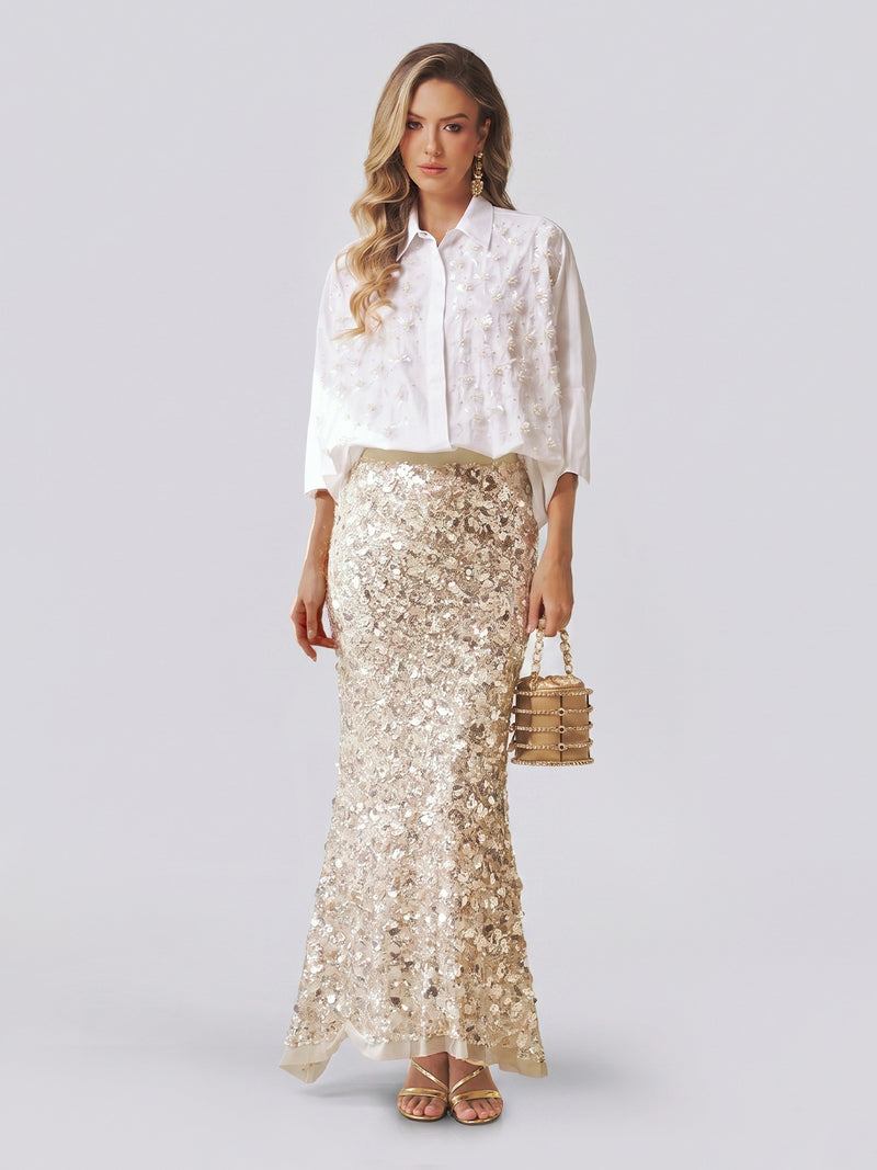 Dressed in Social Queen’s Pearl-Embellished Floral Blouse and a long, sequined beige skirt, the woman holds a small handbag while standing against a plain light background.