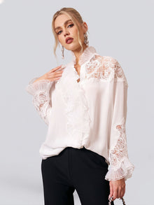 A woman wears the Social Queen Ruffled Lace Blouse with Asymmetric Hem, featuring pleated organza ruffles and long sleeves, paired with black pants, standing against a plain light background.