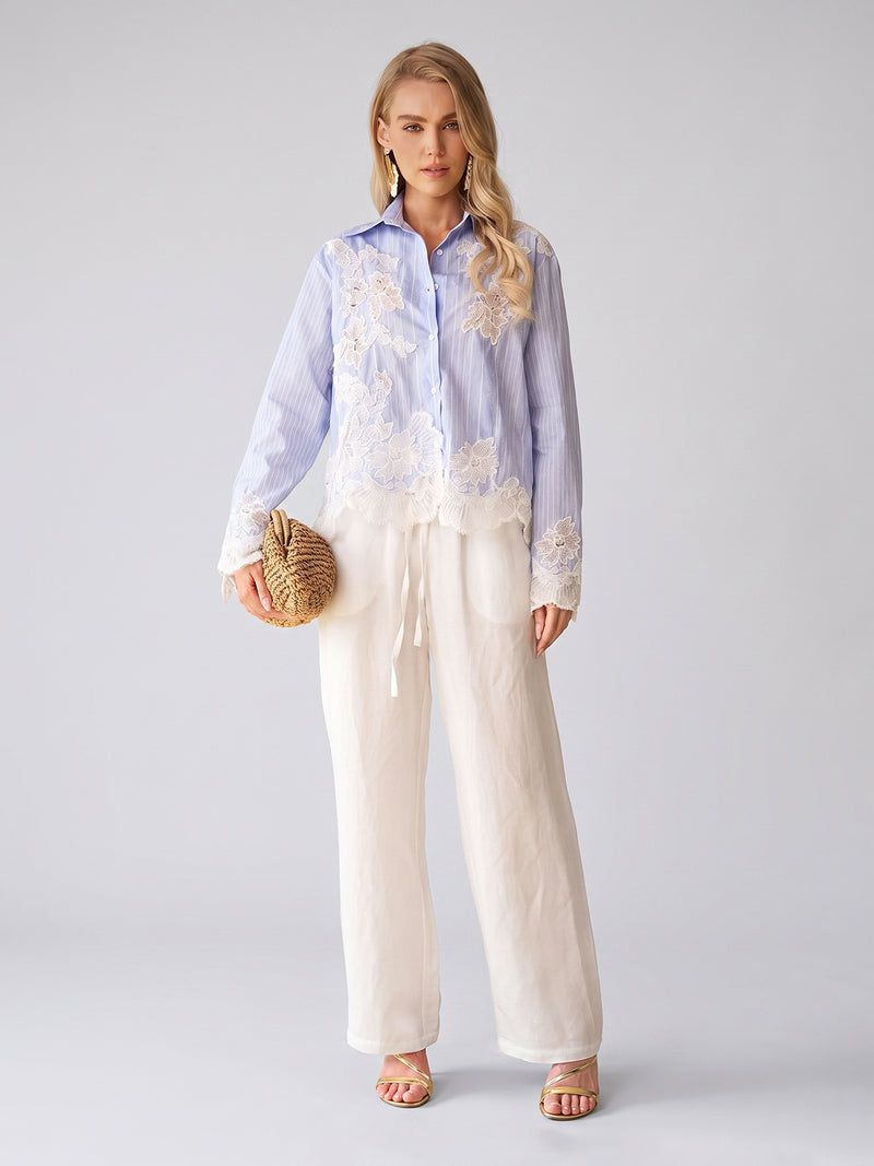 A woman stands against a gray background wearing the Social Queen Lace Panel Striped Cotton Blouse paired with white wide-leg pants, holding a woven clutch, and strappy heels.