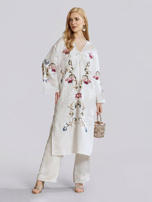 A woman models the Social Queen Botanical Embroidered Linen Top with matching pants, showcasing intricate botanical-inspired embroidery. She holds a small beaded handbag and stands against a plain light background.