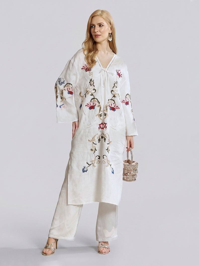A woman models the Social Queen Botanical Embroidered Linen Top with matching pants, showcasing intricate botanical-inspired embroidery. She holds a small beaded handbag and stands against a plain light background.