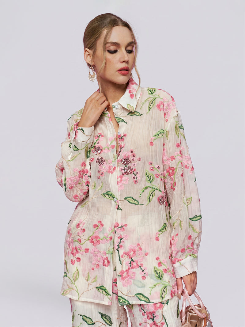 A woman wearing the Social Queen Textured Embroidered Beaded Shirt, featuring sheer long sleeves and pink floral embroidery, holds a pink metallic handbag against a plain light background.