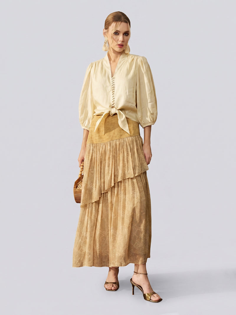 A woman stands against a plain light background, wearing the Social Queen Tie-Front Ramie V-Neck Blouse with puff sleeves and a tie-front hem, paired with a tan layered maxi skirt, gold heeled sandals, and holding a small wooden handbag.