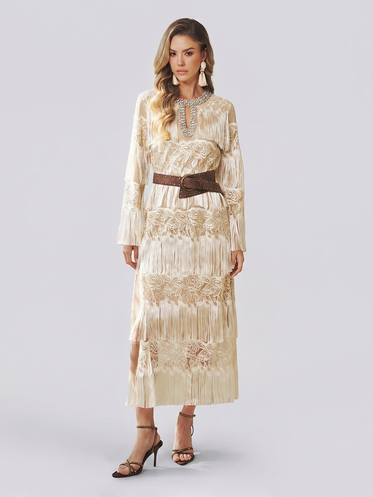 A woman models the Social Queen Embroidered Fringe Lace Crystal-Trimmed Dress—a long cream gown featuring delicate hand-embroidered openwork, lace details, and fringe accents—cinched at the waist with a wide brown belt and styled with a statement necklace and strappy heels against a plain light background.