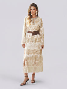 A woman models the Social Queen Embroidered Fringe Lace Crystal-Trimmed Dress—a long cream gown featuring delicate hand-embroidered openwork, lace details, and fringe accents—cinched at the waist with a wide brown belt and styled with a statement necklace and strappy heels against a plain light background.