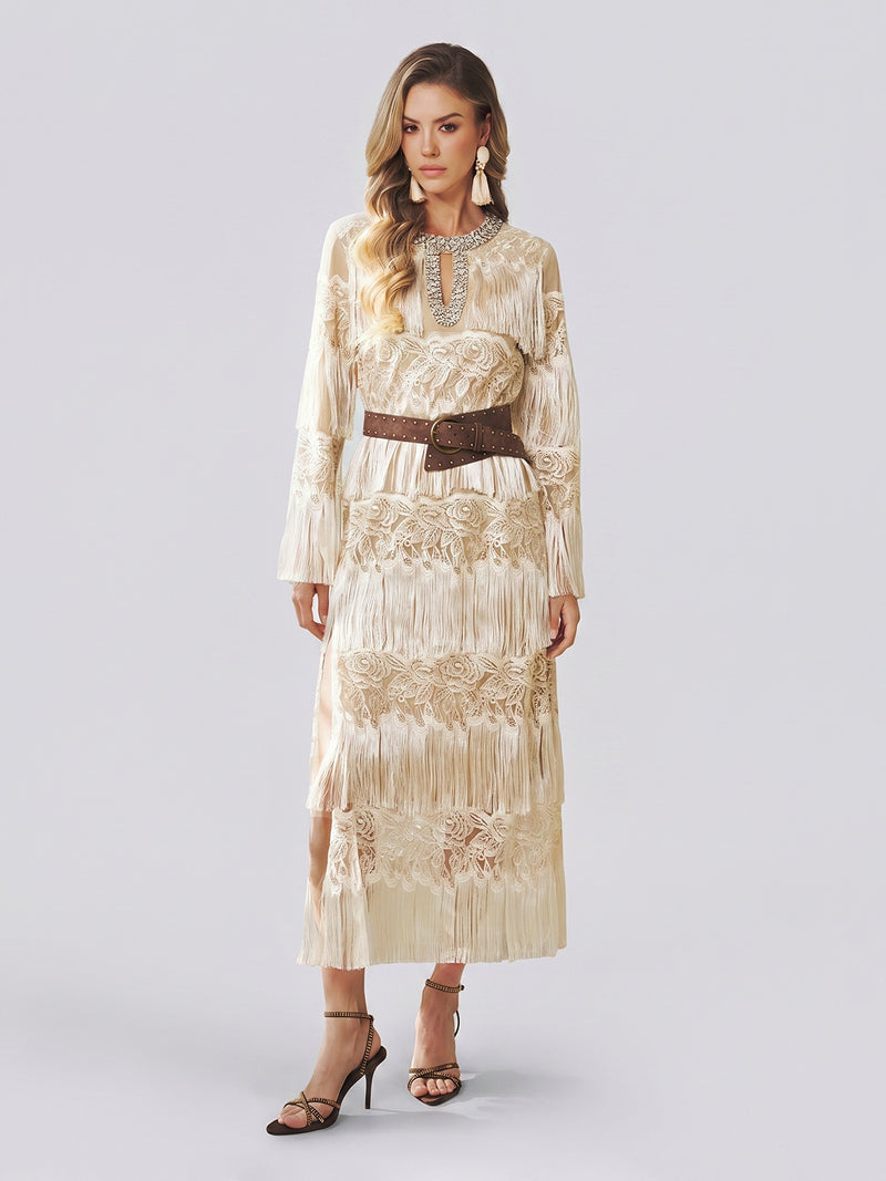 A woman models the Social Queen Embroidered Fringe Lace Crystal-Trimmed Dress—a long cream gown featuring delicate hand-embroidered openwork, lace details, and fringe accents—cinched at the waist with a wide brown belt and styled with a statement necklace and strappy heels against a plain light background.