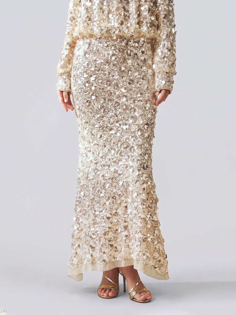 A person poses against a plain background wearing the Social Queen Beaded Sequin Mermaid Elastic Skirt, a floor-length beige skirt featuring allover sequins and a sheer overlay, paired with gold high-heeled sandals.