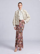 A model wears a light-colored Floral Appliqué Embroidered Jacket by Social Queen over a white ruffled blouse and a floor-length pink skirt with floral embroidery and beadwork.