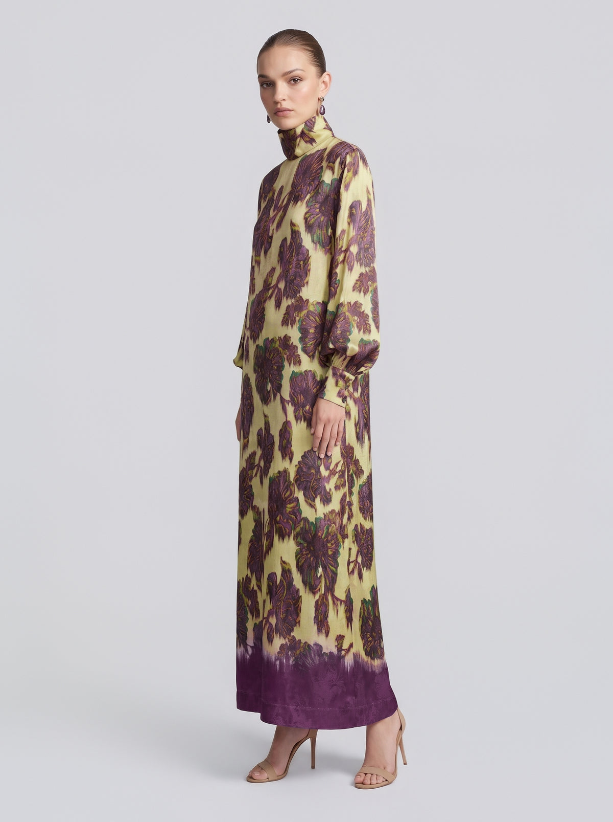 A woman stands in profile against a plain background, wearing the Social Queen Art Floral Jacquard Maxi Dress—a long-sleeved, floor-length gown featuring a yellow and purple floral pattern.