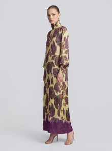 A woman stands in profile against a plain background, wearing the Social Queen Art Floral Jacquard Maxi Dress—a long-sleeved, floor-length gown featuring a yellow and purple floral pattern.