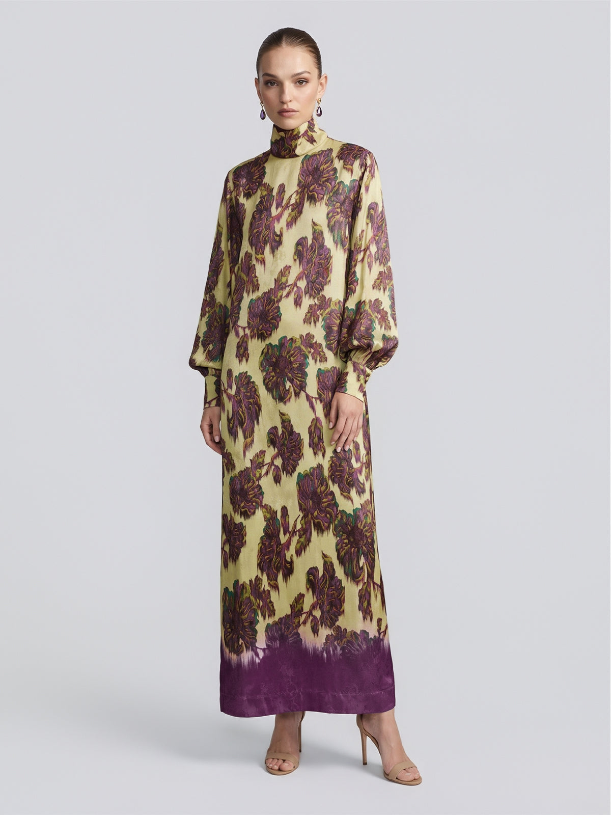 A woman stands against a plain, light background, wearing the Social Queen Art Floral Jacquard Maxi Dress—a long-sleeve, high-neck maxi dress in viscose jacquard featuring a purple floral pattern on a yellow background—paired with open-toe heels.