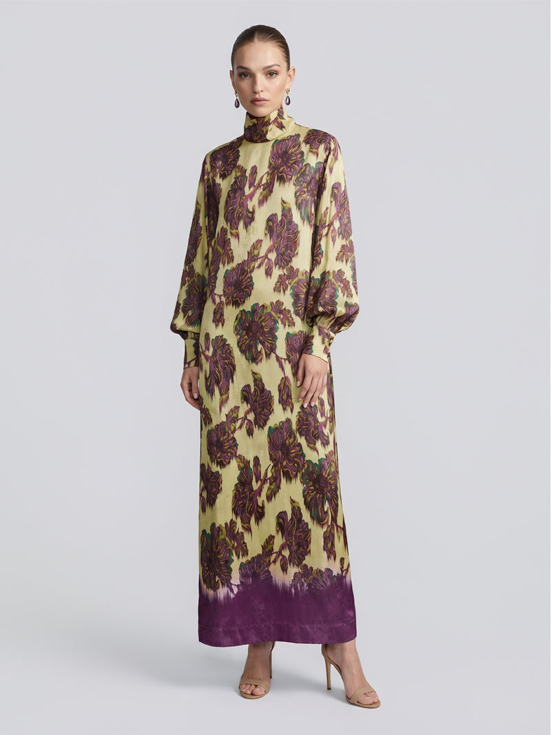 A woman stands against a plain, light background, wearing the Social Queen Art Floral Jacquard Maxi Dress—a long-sleeve, high-neck maxi dress in viscose jacquard featuring a purple floral pattern on a yellow background—paired with open-toe heels.