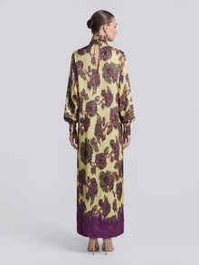 A woman with her hair in a bun models the Social Queen Art Floral Jacquard Maxi Dress, a long-sleeved, floor-length piece featuring purple accents and crafted from luxurious viscose jacquard, shown from the back against a plain light background.
