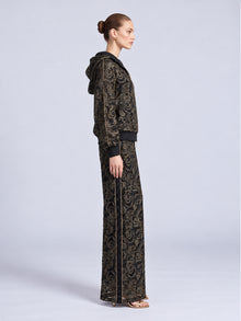 A side view of a person wearing a Social Queen Paisley Embroidered Lace Hooded Jacket in black and gold with matching wide-leg trousers featuring a black side stripe.