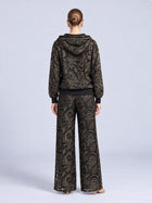 A model from behind wearing a black hooded jacket and matching Paisley Embroidered Lace Wide-Leg Pants, both featuring gold paisley embroidery on cutout lace fabric.