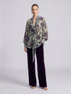 A model wears a sheer, long-sleeved blouse with a green and purple floral pattern and dark purple Social Queen Velvet Wide-Leg Pants.