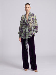 A model wears a sheer, long-sleeved blouse with a green and purple floral pattern and dark purple Social Queen Velvet Wide-Leg Pants.