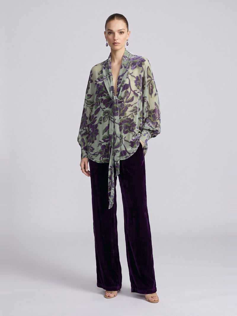 A model wears a sheer, long-sleeved blouse with a green and purple floral pattern and dark purple Social Queen Velvet Wide-Leg Pants.
