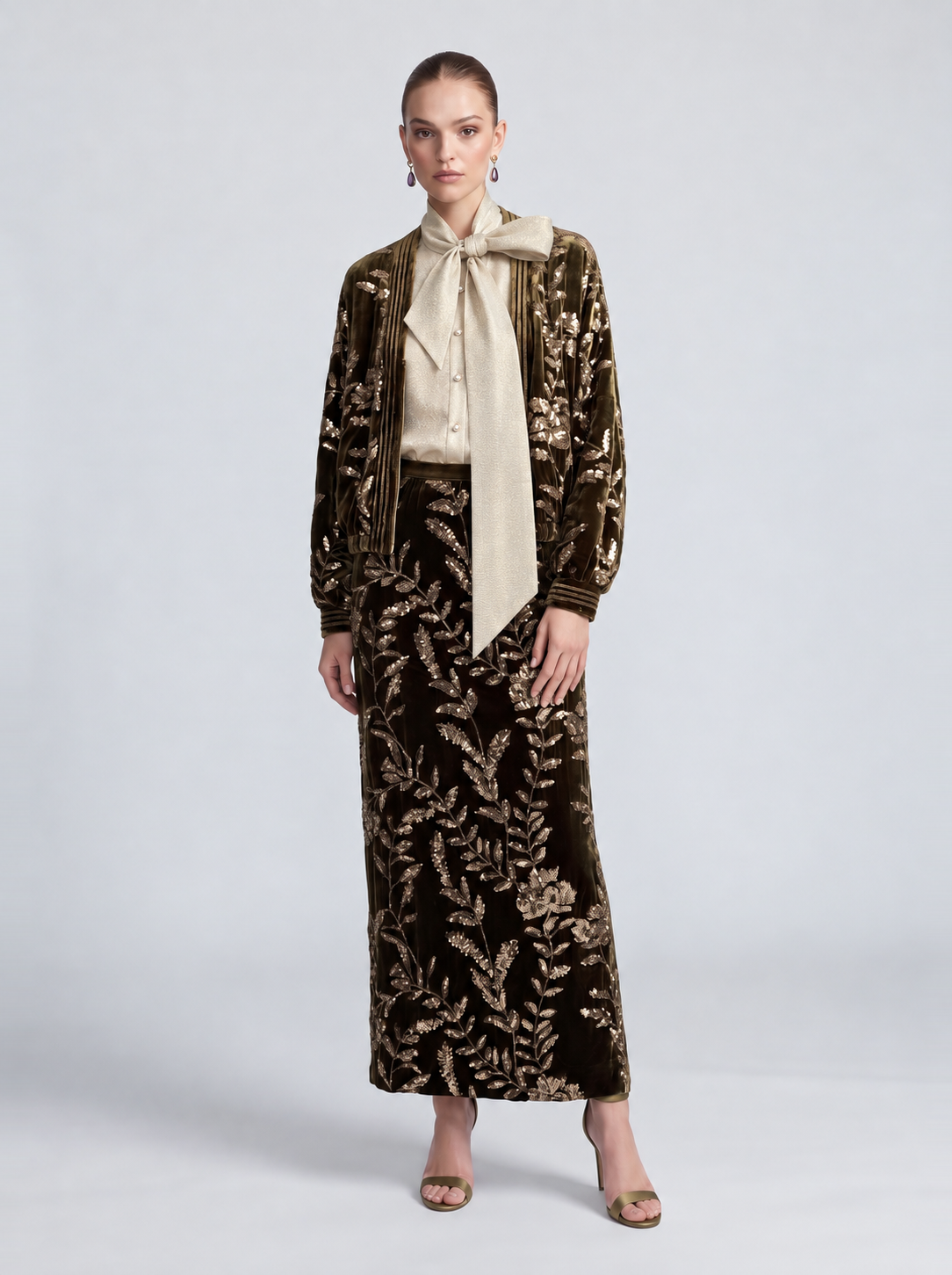 A model wearing a Social Queen Sequin Embroidered Velvet Midi Skirt in dark olive with gold-tone floral sequins, a matching zip-up jacket, and a cream-colored pussy-bow blouse.