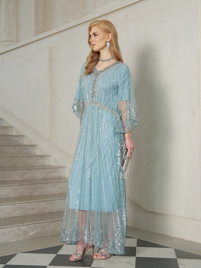 A woman in the Airy Beaded Batwing Tulle Dress by Social Queen stands indoors near stairs, holding a clutch. She pairs the look with silver heels, a necklace, earrings, and straight side-parted hair.