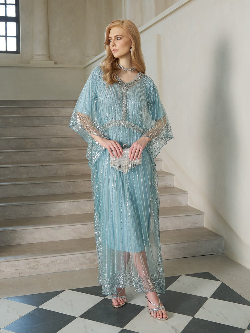 A woman in the Social Queen Airy Beaded Batwing Tulle Dress stands on a staircase, looking to the side. She pairs the light blue, sheer gown with silver sandals and jewelry, her long hair styled in loose waves.
