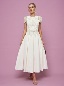 A woman poses against a pink background in the Social Queen Appliqué Pearl Waffle Mesh Dress—white, knee-length, with an A-line silhouette and short sleeves—paired with silver sandals and a small metallic handbag.