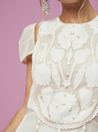 A close-up of a woman in the Social Queen Appliqué Pearl Waffle Mesh Dress, featuring floral patterns and pearl embellishments, set against a pink background with part of her face and a stylish earring visible.