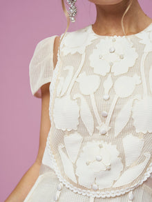 A close-up of a woman in the Social Queen Appliqué Pearl Waffle Mesh Dress, featuring floral patterns and pearl embellishments, set against a pink background with part of her face and a stylish earring visible.