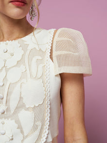 Close-up of a woman in the Social Queen Appliqué Pearl Waffle Mesh Dress, showing its floral patterns, mesh puff sleeves, and elegant A-line silhouette. Only her lower face, shoulder, and part of an earring are visible against a pink background.