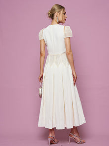 A woman stands against a pink background, facing away, wearing the Social Queen Appliqué Pearl Waffle Mesh Dress—an A-line white midi dress with sheer short sleeves—paired with a small silver bag and strappy high heels.