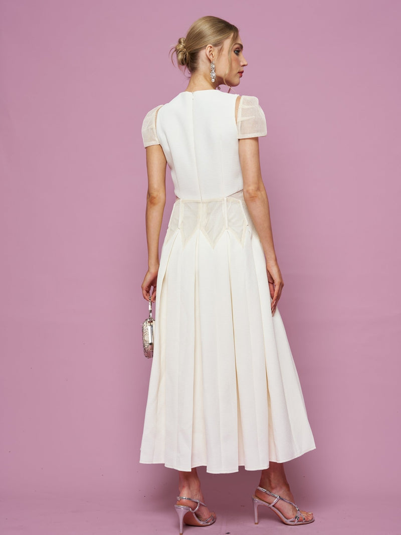 A woman stands against a pink background, facing away, wearing the Social Queen Appliqué Pearl Waffle Mesh Dress—an A-line white midi dress with sheer short sleeves—paired with a small silver bag and strappy high heels.
