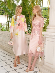 Two women stand on a tiled terrace with green foliage behind. One wears the Social Queen Aquarelle Blooms 100% Mulberry Silk Dress, while the other dons a pink lace dress. Both accessorize with decorative handbags and sandals.