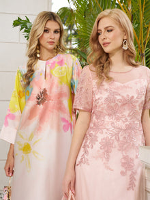 Two women with long blonde hair stand together. One wears the Social Queen Aquarelle Blooms 100% Mulberry Silk Dress, featuring a watercolor floral print; the other wears a fitted pink floral embroidered wedding guest dress. Green plants are behind them.