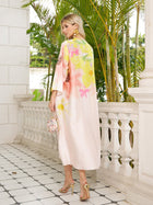 On a tiled balcony, a woman in Social Queen’s Aquarelle Blooms 100% Mulberry Silk Dress and gold heels holds a small floral handbag, with palm plants behind her.