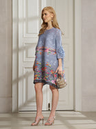 A woman indoors wears the Social Queen Art-Print Floral-Appliqué Pleated Dress with ruffled sleeves, carries a textured handbag, lets her hair down, and completes her look with strappy heels.