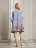 A woman with long blonde hair stands indoors, facing away, wearing the Social Queen Art-Print Floral-Appliqué Pleated Dress in light blue, styled with white heeled sandals.