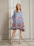 A woman indoors wears the Social Queen Art-Print Floral-Appliqué Pleated Dress, featuring ruffled sleeves, styled with silver heels and a small handbag.
