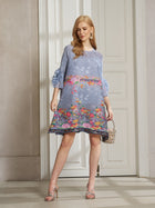 A woman indoors wears the Social Queen Art-Print Floral-Appliqué Pleated Dress, featuring ruffled sleeves, styled with silver heels and a small handbag.