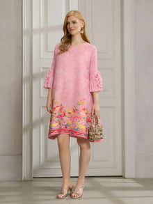 A woman stands indoors wearing the Social Queen Art-Print Floral-Appliqué Pleated Dress in light pink with ruffled sleeves, paired with silver sandals and a small metallic handbag.