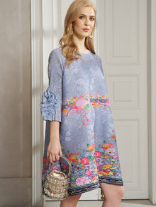 A woman stands indoors holding a small metallic handbag while wearing the Social Queen Art-Print Floral-Appliqué Pleated Dress with ruffled sleeves.