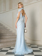 A woman stands on a checkered floor near a staircase, wearing the Social Queen Artisanal Beaded Fringe High-Neck Gown in light blue, holding a small handbag and looking over her shoulder.
