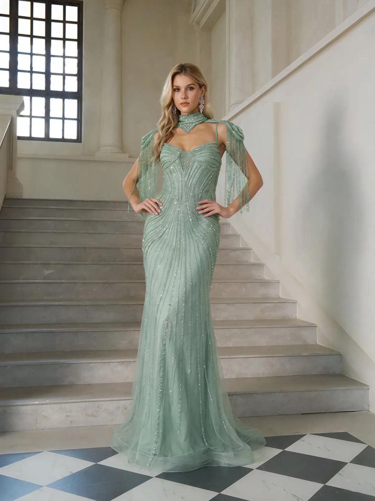 A woman stands on indoor stairs wearing the Artisanal Beaded Fringe High-Neck Gown by Social Queen, paired with statement earrings—an exquisite choice for any formal event.