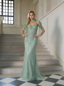 A woman stands on indoor stairs wearing the Artisanal Beaded Fringe High-Neck Gown by Social Queen, paired with statement earrings—an exquisite choice for any formal event.