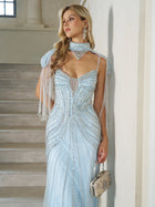 A woman stands by a staircase in the Social Queen Artisanal Beaded Fringe High-Neck Gown, holding a silver clutch and wearing statement earrings and a choker—a striking choice for any formal occasion.