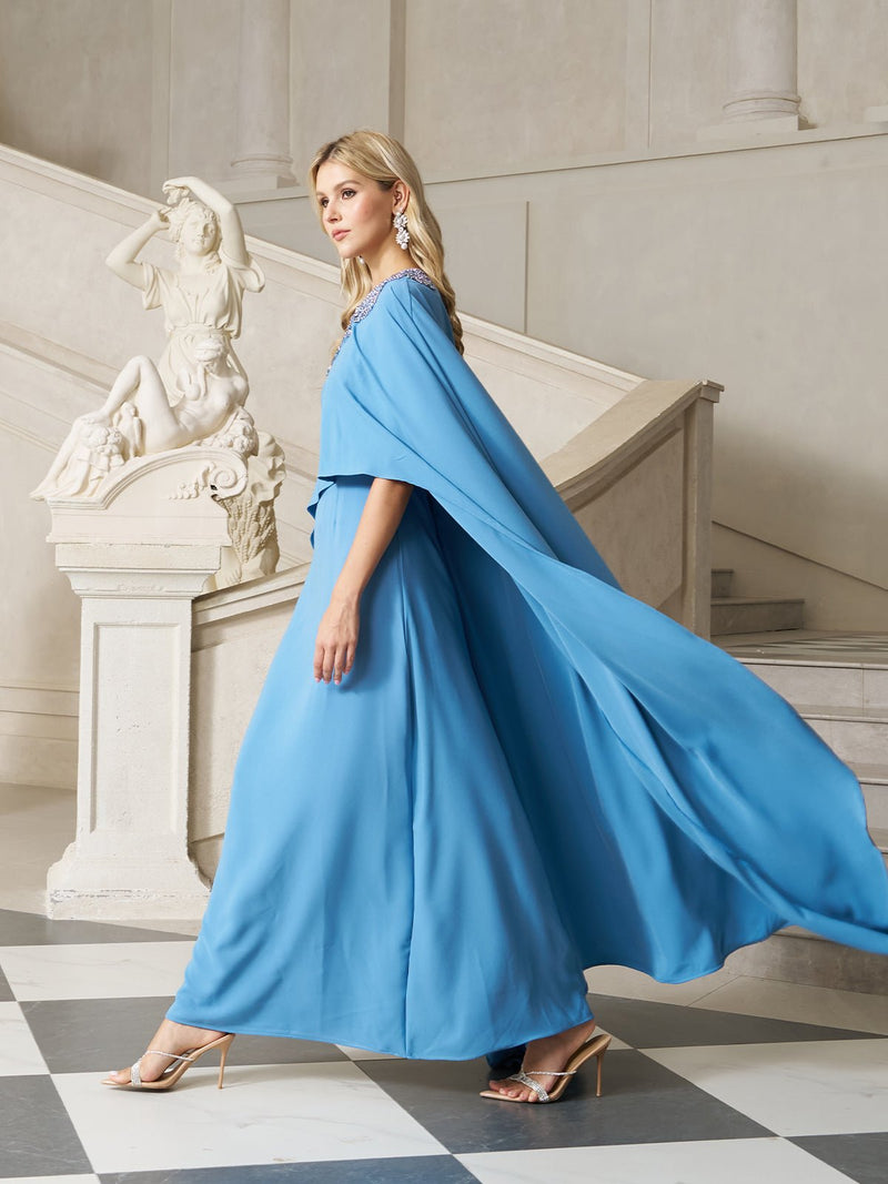 A formal wedding guest in Social Queen’s Artisanal Embellished Cape Dress and heels passes a white statue in a classical interior with stairs and checkered flooring.