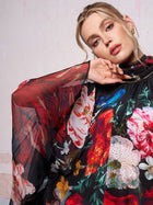 A woman models the Social Queen Artistic Floral Print Ruffle Dress, featuring sheer fabric with bold, colorful patterns, against a light-colored wall.