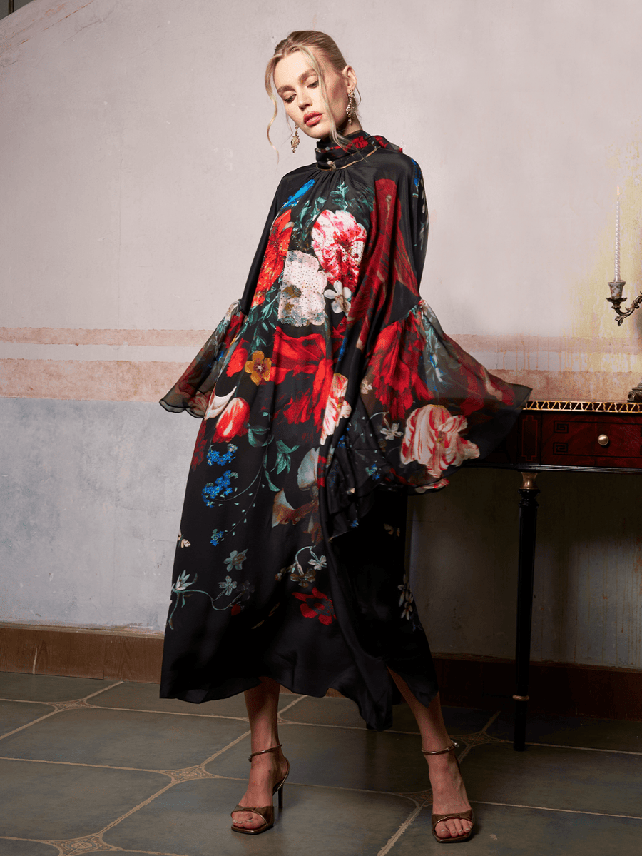 A woman poses indoors in the Social Queen Artistic Floral Print Ruffle Dress, featuring a long, flowing silhouette and vibrant floral patterns, standing beside a table with a single candleholder.