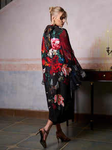 A woman in the Social Queen Artistic Floral Print Ruffle Dress and gold heels stands indoors, facing away from the camera near a wooden table with a brass candlestick.